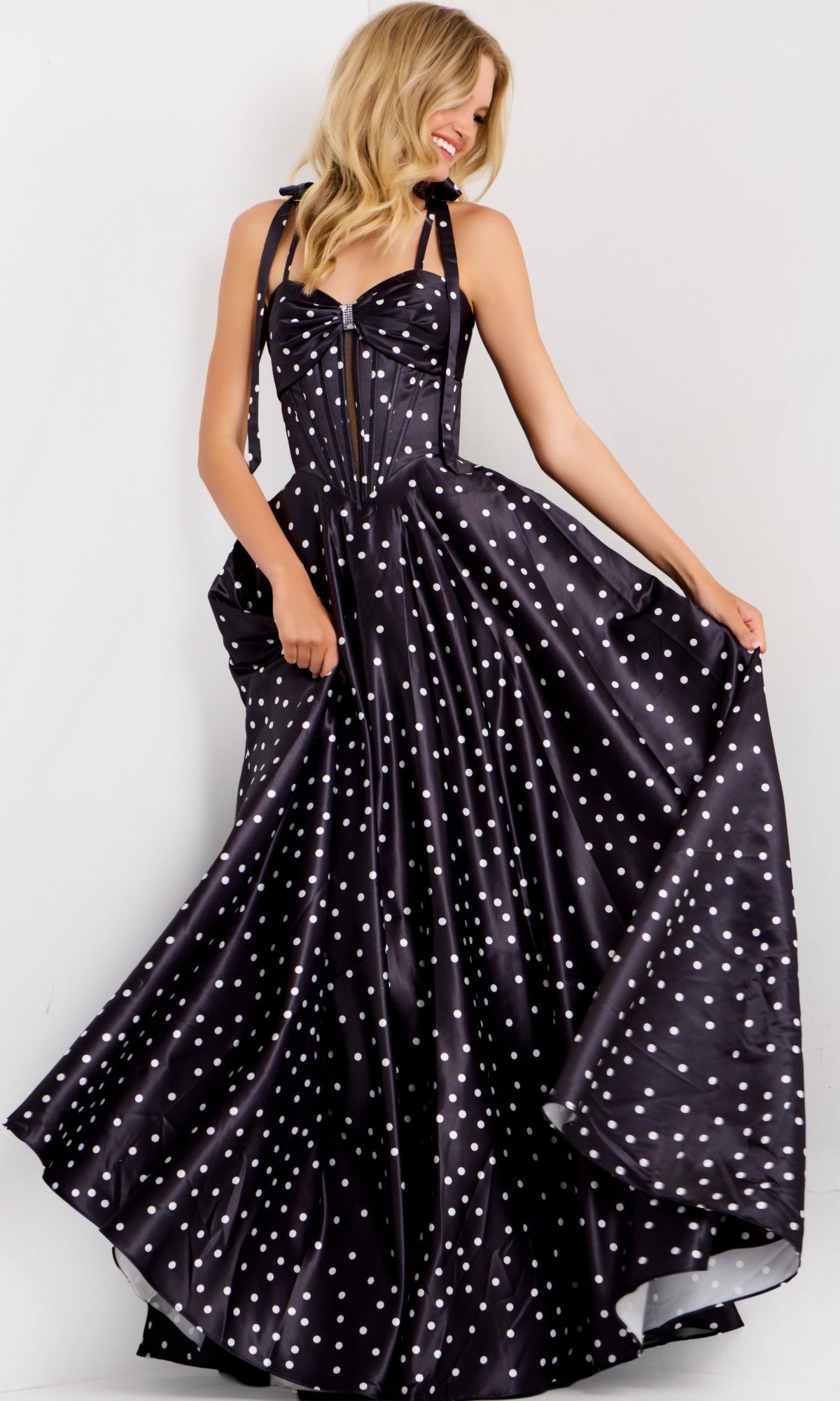 A woman poses against a plain background in the Jovani 49774 long prom dress, featuring black satin with polka dots and a flowing a-line skirt, holding out the sides of her skirt.